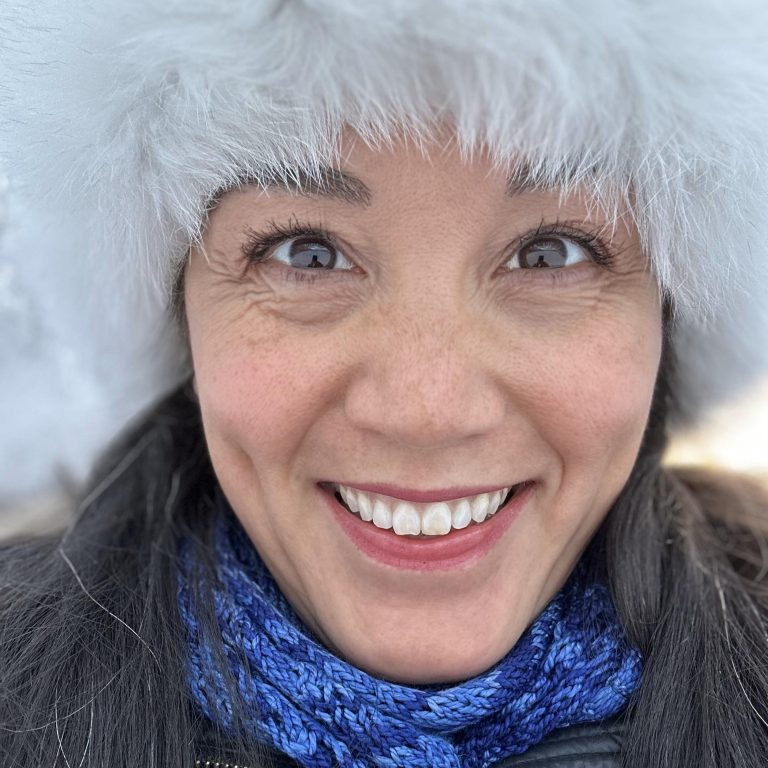 Close-up of Chris Brewer in winter smiling and looking directly into the camera. She is wearing a white fur hat and blue scarf.